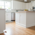 Sunlit modern kitchen with white cabinets, kitchen island, stainless steel refrigerator, and hardwood floors, showing a cockroach on the floor near the cabinet base.